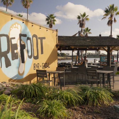 A sunny beachside bar with palm trees, patio tables, and a &ldquo;BA RBE Q&rdquo; mural on a yellow wall.