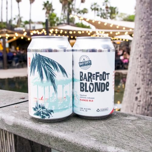 Two Bay City &ldquo;Barefoot Blonde&rdquo; beer cans sit on a stone ledge at an outdoor festival with string lights.