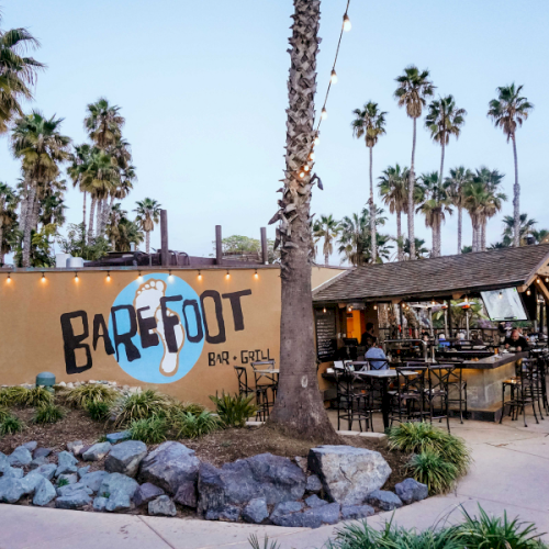 An outdoor &ldquo;Barefoot&rdquo; bar and grill with string lights, patio seating, and tall palm trees in the background.