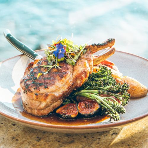 A seaside plate of grilled pork chops with roasted veg, broccoli, and microgreens, topped with edible flowers and a knife.