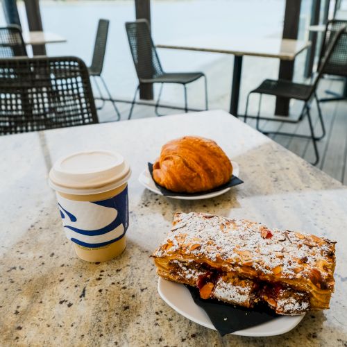 Coffee and a pastry on a patio table by the water, with lounge chairs in the background.