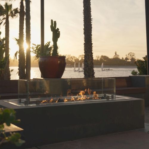 An outdoor beachfront fire pit burning at sunset, framed by palm trees and string lights over calm water and boats in the distance.