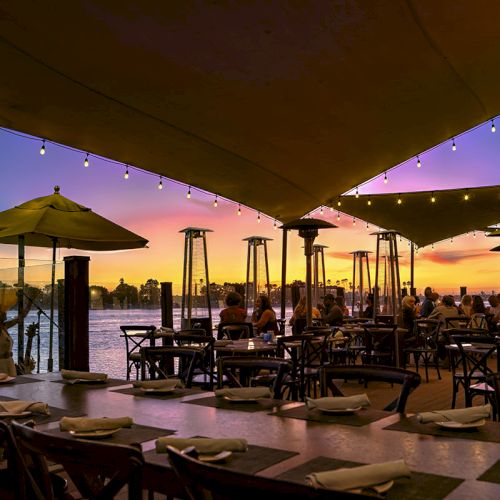 Outdoor lakeside dining at sunset with candle-lit string lights, patio tables, and umbrellas under a covered deck.