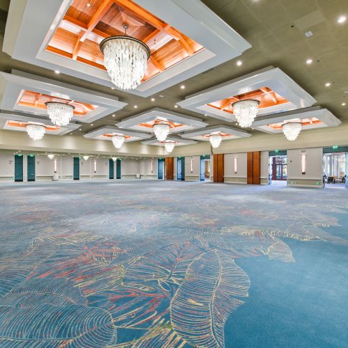 A spacious conference hall with a blue carpet, wood ceiling beams, and multiple crystal chandeliers hanging overhead, topped with skylit ceiling panels.
