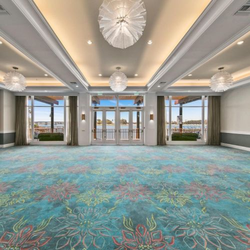 An elegant indoor hall with patterned carpet, curtains, and large windows, lit by hanging chandeliers and recessed lights.