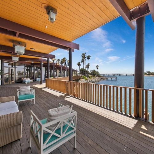 A sunny waterfront porch with wood railings, patio seating, and a view of calm ocean water and palm trees.