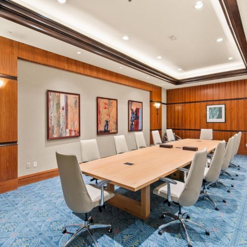 A modern conference room with a large wooden table, swivel chairs, blue patterned carpet, framed art, and warm lighting.