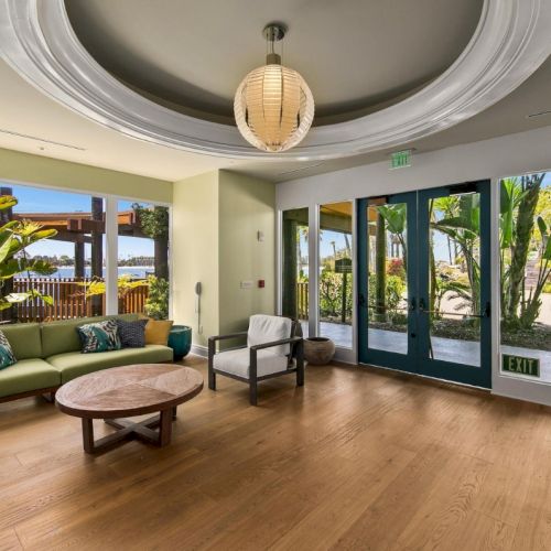 A bright modern lounge with a green sofa, round wooden coffee table, tropical plants, and glass doors by an &ldquo;EXIT&rdquo; sign.