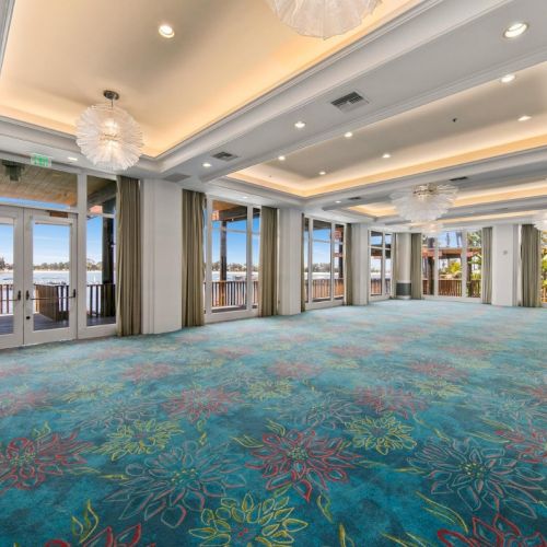 Bright hotel event hall with blue patterned carpet, white chandeliers, and large windows overlooking the waterfront.