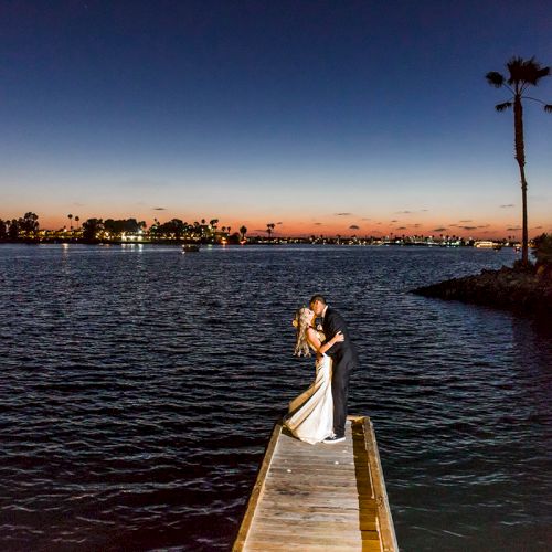 A couple kisses on a wooden dock at sunset over a lake, with palm trees and city lights in the distance.