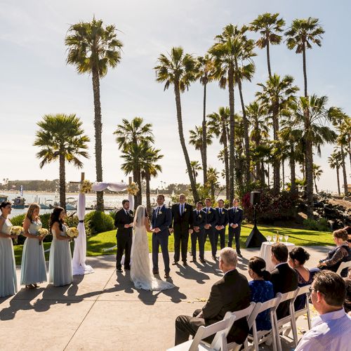 Outdoor wedding ceremony by the ocean with bridesmaids and groomsmen in suits, guests seated, palm trees overhead.