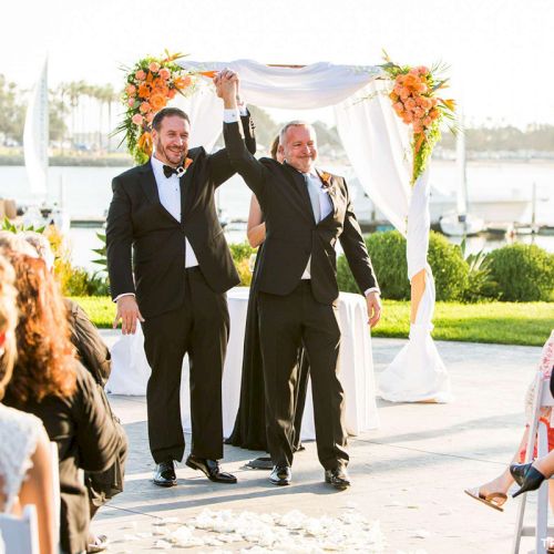 An outdoor wedding ceremony by the water as two men in tuxedos walk forward under a floral arch while guests watch.