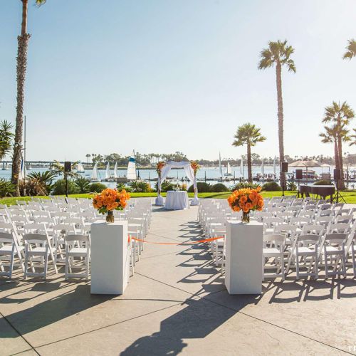 Outdoor wedding setup with rows of white chairs, orange flower arrangements, tall palm trees, and a view of the ocean.