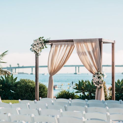 A beachfront wedding ceremony setup: white chairs facing a floral draped arch with the ocean and bridge in the background.