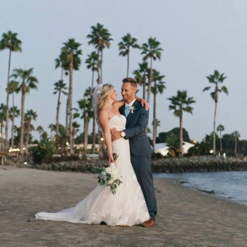 A bride and groom in wedding attire pose on a palm-lined beach at dusk, holding a bouquet, with the ocean beside them.