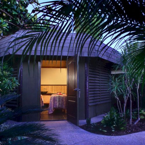A tropical garden courtyard with an open archway revealing a warmly lit room, a covered table, and lush surrounding plants, at dusk.