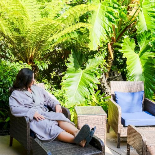 A person relaxes on an outdoor lounge chair in a lush tropical garden surrounded by ferns, banana leaves, and potted plants.