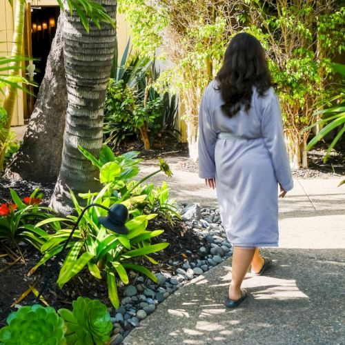 A woman in a light blue robe walks along a sunny stone path in a lush tropical garden lined with plants and a palm tree.