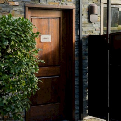 A wooden door set in a stone wall, with a mailbox, leafy shrubbery, and a dark outdoor grill/cabinet nearby.