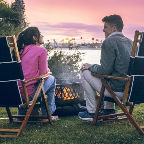 Two people sit in deck chairs by a small fire on the grass, watching the sunset over a lake.