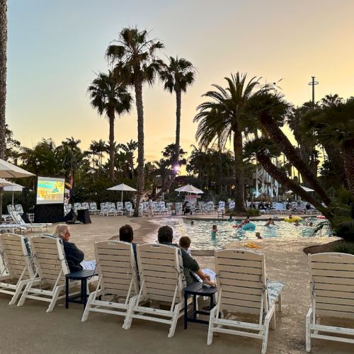 Sunset poolside at a resort with palm trees, lounge chairs, umbrellas, and swimmers in the water.