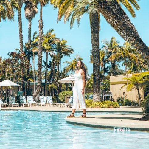A woman stands by a sunny resort pool surrounded by tall palm trees, soaking up tropical vibes.