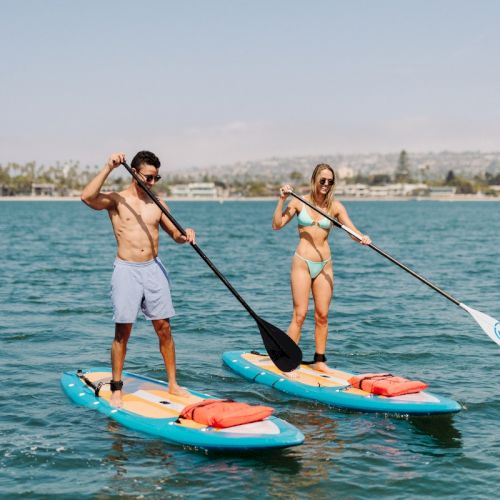 Two people paddleboarding in the ocean, smiling while rowing with paddles against a sunny coastal backdrop.