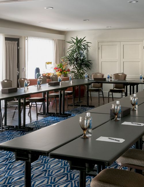 A bright meeting room with long rectangular tables, cushioned chairs, place settings, and flowers by a wall.