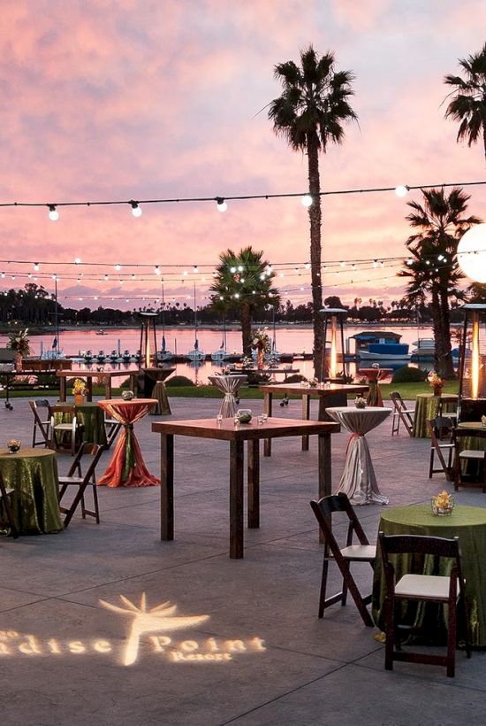 An elegant outdoor party setup with string lights, decorated tables, and palm trees by a marina at sunset, with a glowing bar.