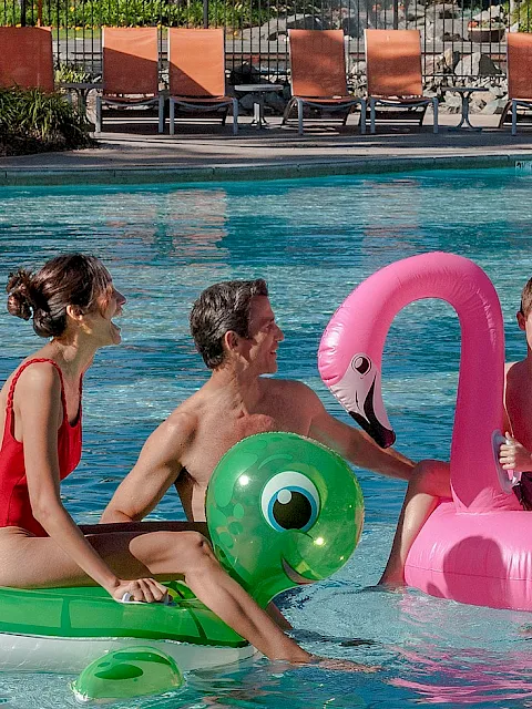 Kids play in a sunny outdoor pool with colorful inflatable rings and a green-and-pink float.