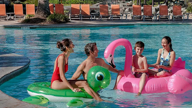 Kids play in a sunny outdoor pool with colorful inflatable rings and a green-and-pink float.