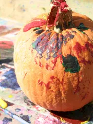 A painted pumpkin sits on an art-painted table, surrounded by colorful brushes and paper scraps, ready for decoration.