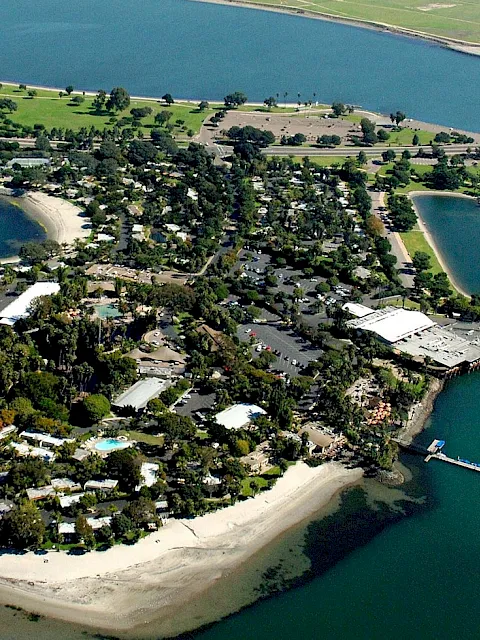 Aerial view of a small coastal island resort with beaches, docks, a bridge, and pools surrounded by blue water.