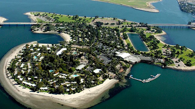 Aerial view of a small coastal island resort with beaches, docks, a bridge, and pools surrounded by blue water.