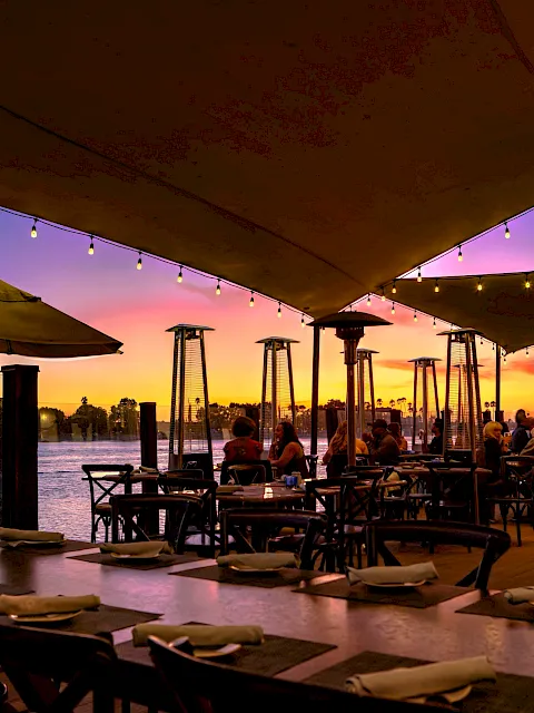 Sunset view of a waterfront restaurant patio with string lights, patio heaters, umbrellas, and diners gathered by the ocean.