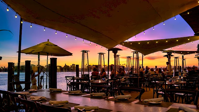 Sunset view of a waterfront restaurant patio with string lights, patio heaters, umbrellas, and diners gathered by the ocean.