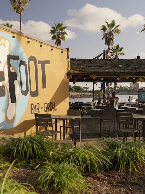 A tropical outdoor bar & grill by the water with palm trees, patio seating, and a big &ldquo;BAR E GRILL&rdquo; sign on a wall.
