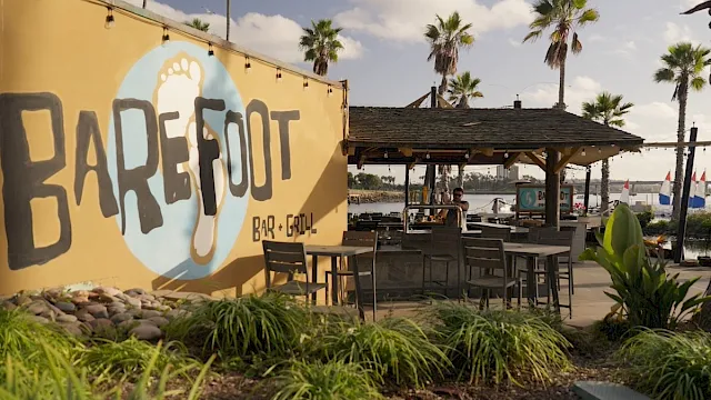 A tropical outdoor bar & grill by the water with palm trees, patio seating, and a big &ldquo;BAR E GRILL&rdquo; sign on a wall.