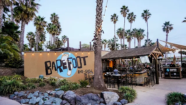 Tropical outdoor bar-grill with string lights, tall palm trees, and the &ldquo;Barefoot&rdquo; sign beside diners.