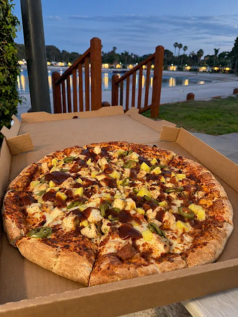A cheese-and-topping pizza in an open cardboard box, topped with pepperoni, onions, and jalape&ntilde;os by a waterfront at dusk.