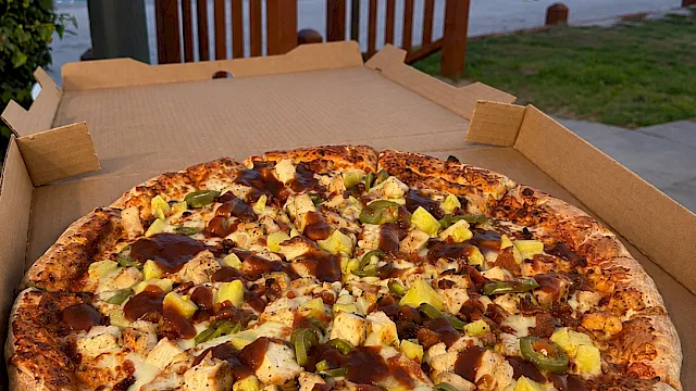 A cheese-and-topping pizza in an open cardboard box, topped with pepperoni, onions, and jalape&ntilde;os by a waterfront at dusk.