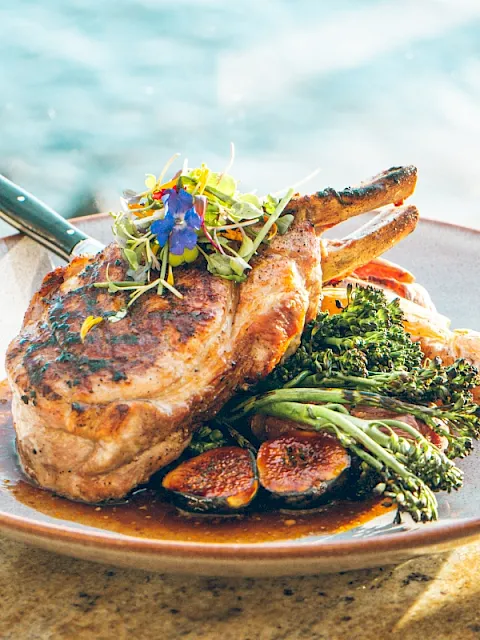 A plate of grilled steak chops topped with microgreens and served with broccoli and roasted vegetables by the ocean.