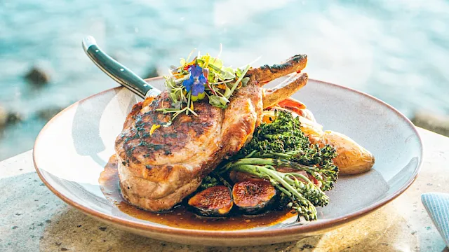 A plate of grilled steak chops topped with microgreens and served with broccoli and roasted vegetables by the ocean.