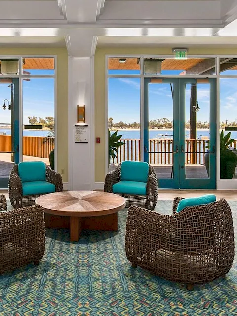 A modern hotel lounge with patterned carpet, wicker chairs, round coffee table, and big doors/windows opening to a seaside view.