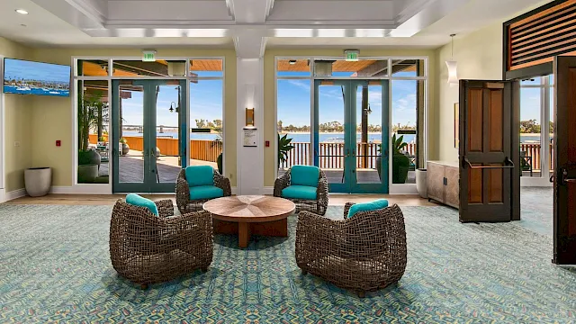 A modern hotel lounge with patterned carpet, wicker chairs, round coffee table, and big doors/windows opening to a seaside view.