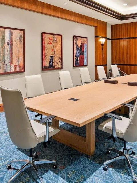 A bright conference room with a large wooden boardroom table, white chairs, framed art on walls, and a blue patterned carpet.