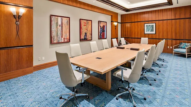 A bright conference room with a large wooden boardroom table, white chairs, framed art on walls, and a blue patterned carpet.