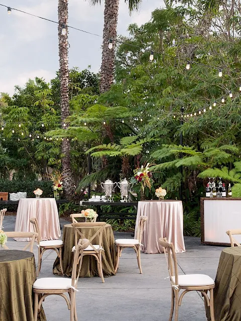 An outdoor wedding reception setup with white covers, flowers, chairs, and a bar table under string lights among palms.