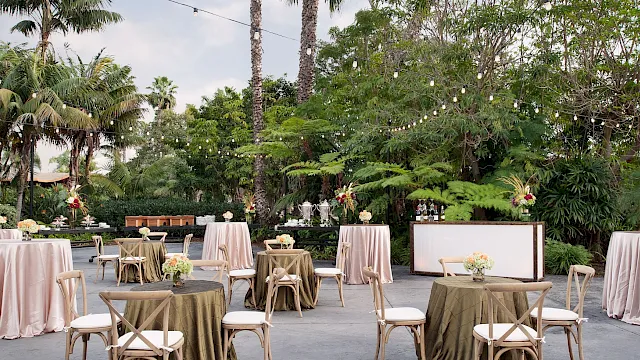 An outdoor wedding reception setup with white covers, flowers, chairs, and a bar table under string lights among palms.