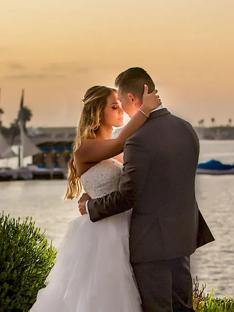 A couple in wedding attire embraces by the waterfront at sunset, with sailboats and palm-lined docks in the background.